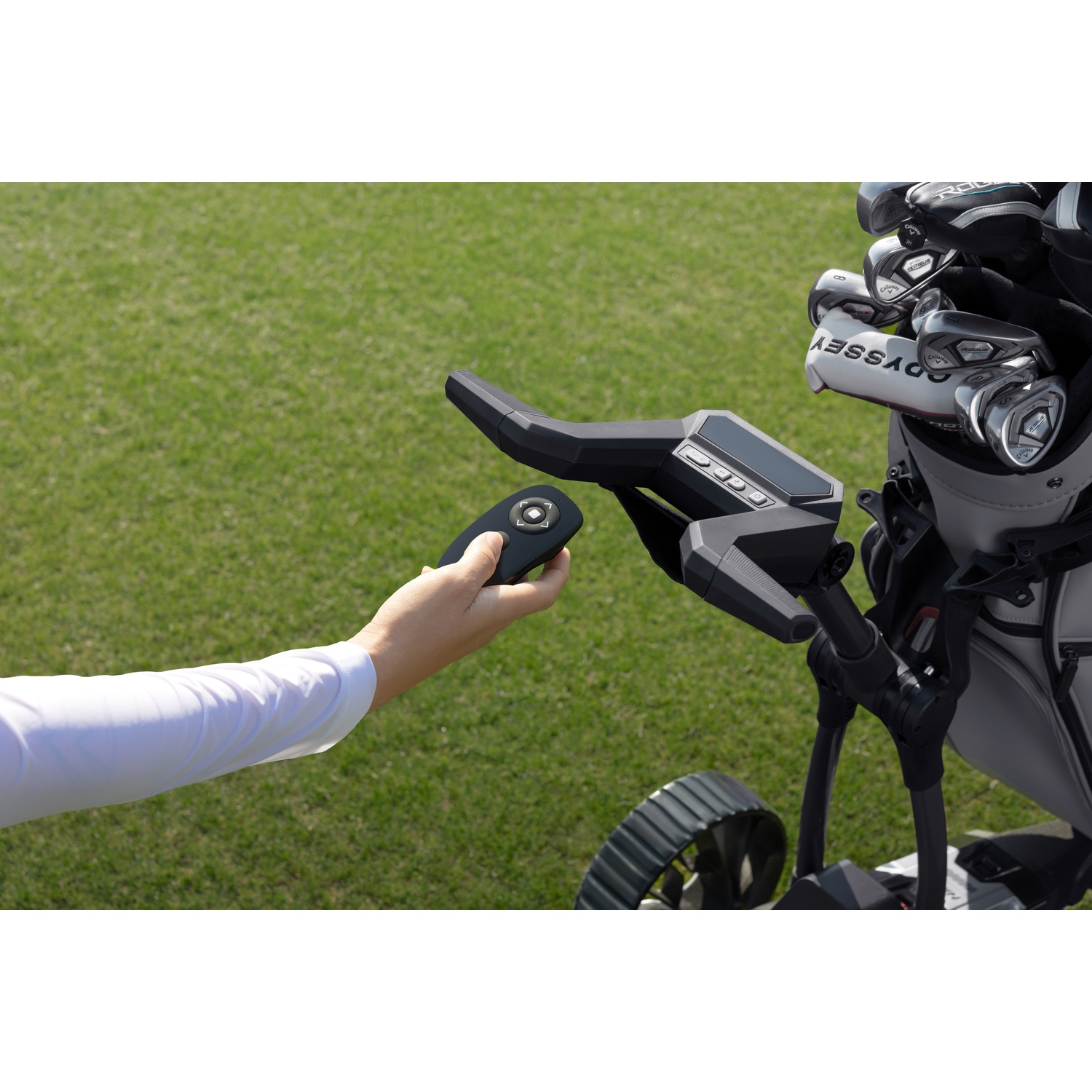 Een hand houdt een afstandsbediening vast naast een golfkar met clubs op gras.
