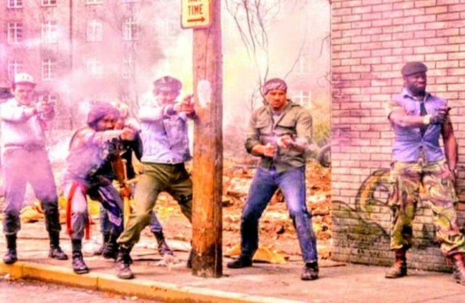 Fünf Männer mit Waffen posieren vor einer Backsteinmauer und Gebäuden.