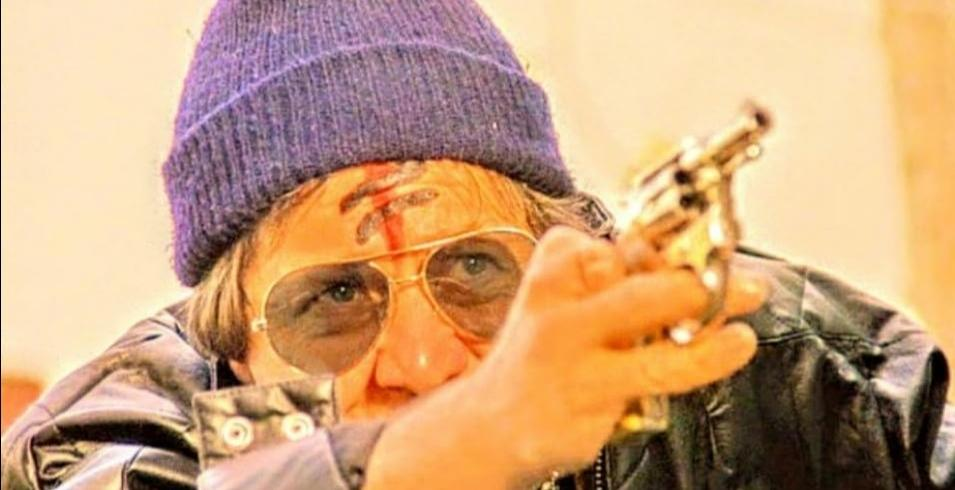 Mann mit lila Mütze, Sonnenbrille und rotem Stirnzeichen hält eine goldene Pistole.