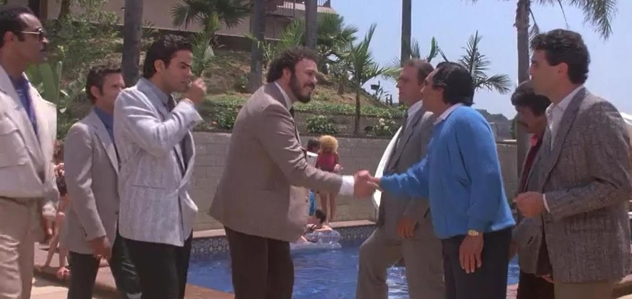 Männer in Anzügen schütteln Hände am Pool, Palmen im Hintergrund.