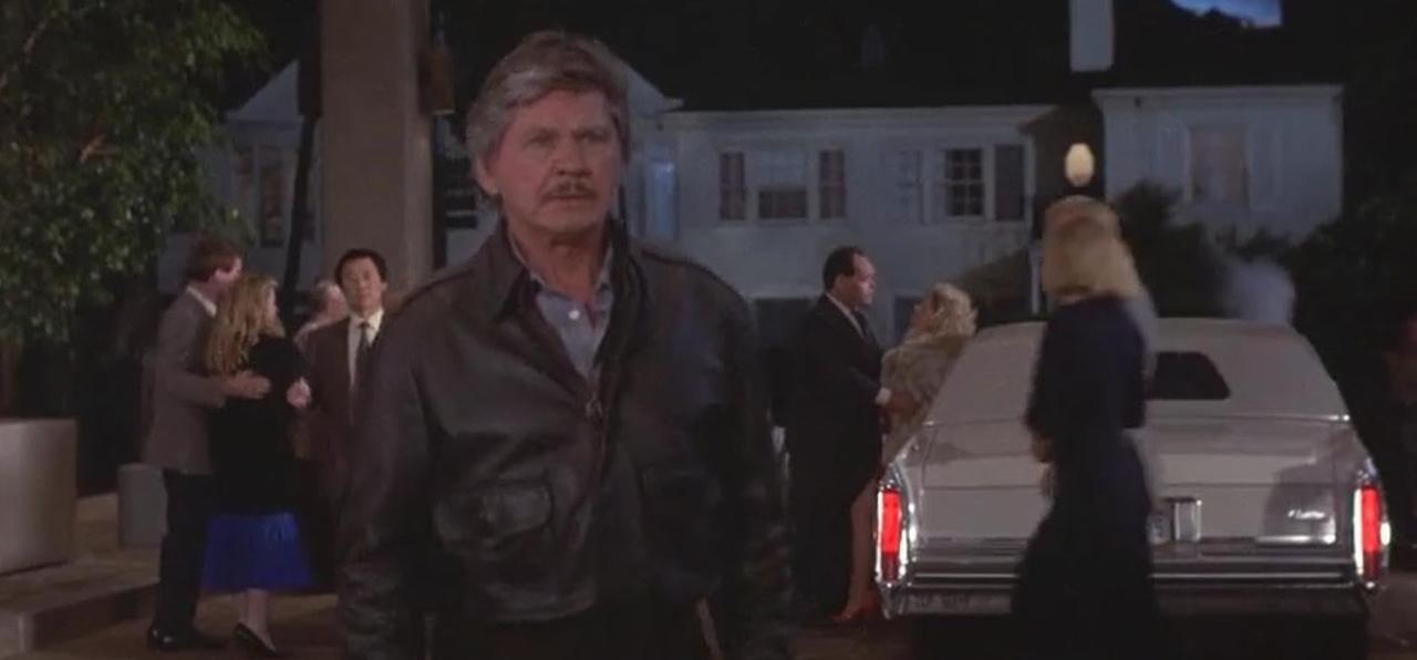 Ein Mann in Lederjacke steht im Vordergrund, im Hintergrund eine weiße Limousine und Haus.
