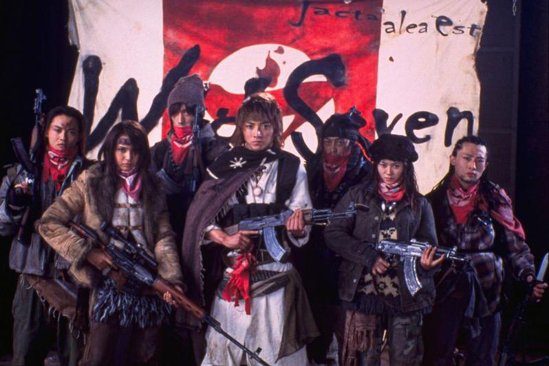 Sieben Personen mit Waffen posieren vor einem Banner.