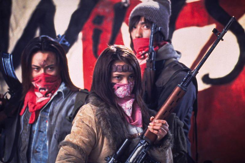 Drei Personen mit Waffen und Bandanas vor einem rot-schwarzen Banner.