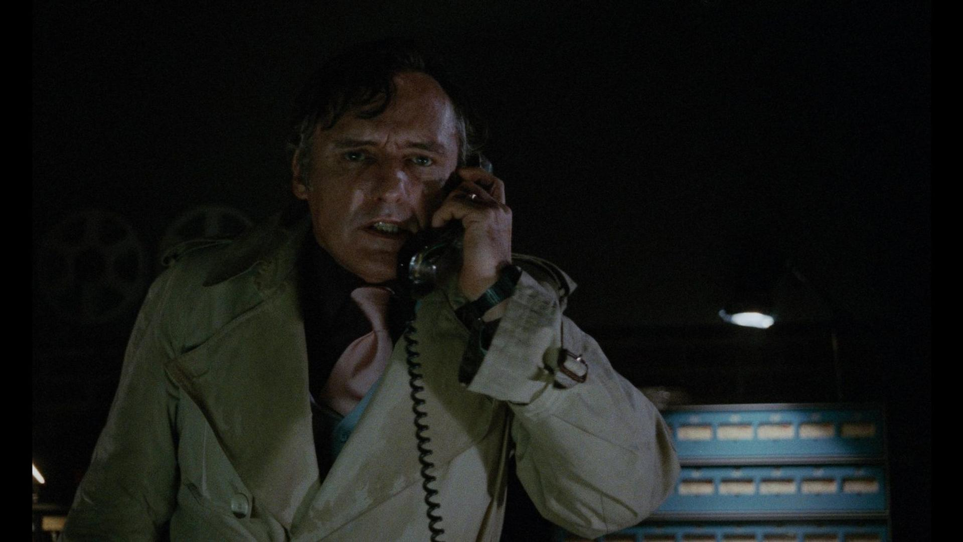 Ein Mann im Trenchcoat telefoniert in einem dunklen Raum mit blinkenden Lichtern.
