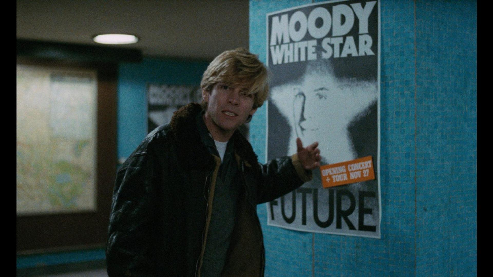 Ein Mann mit blondem Haar steht vor einem Plakat mit der Aufschrift „MOODY WHITE STAR“ und „FUTURE“.