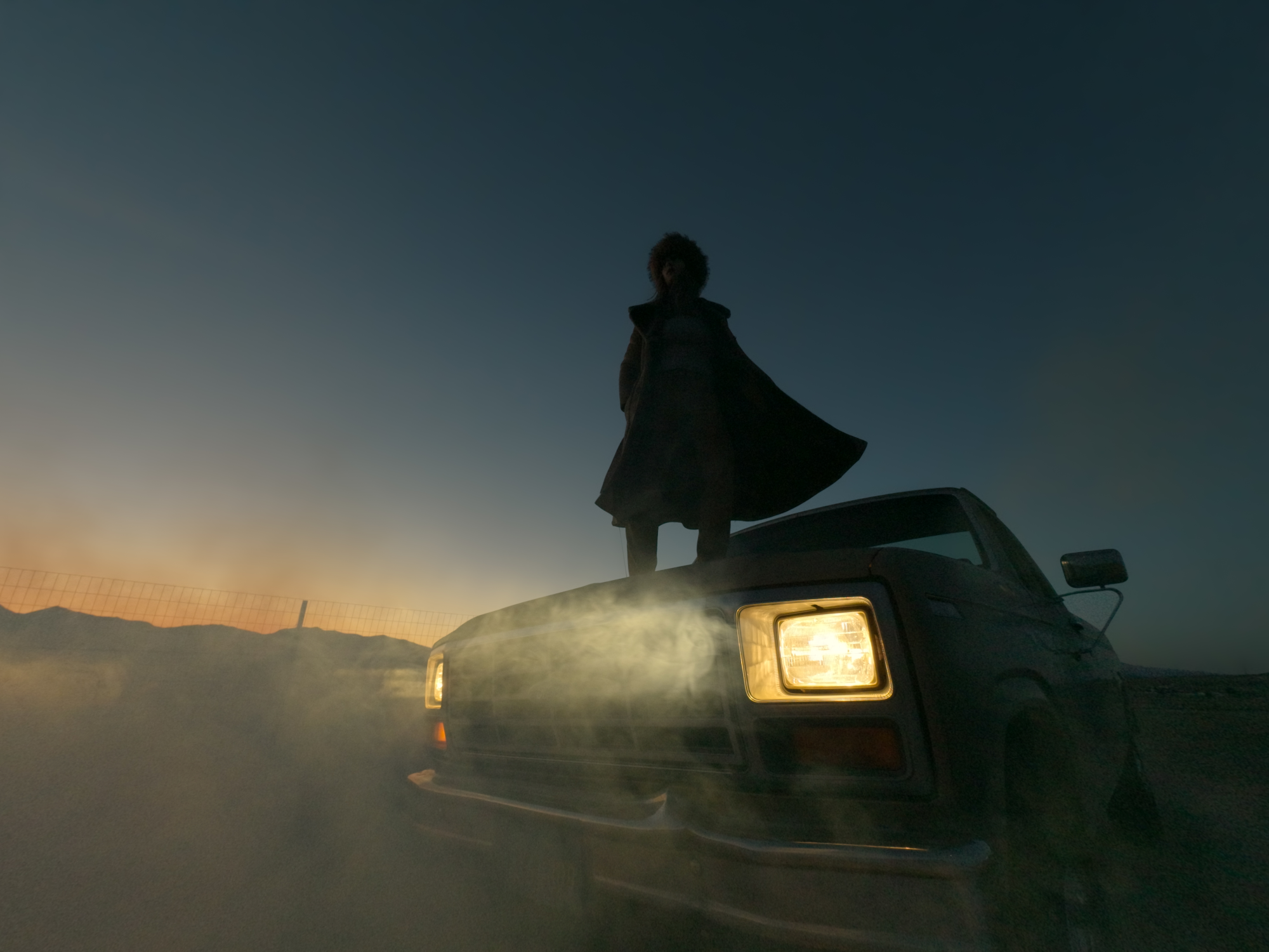 Une personne se tient sur une camionnette avec du brouillard autour des phares au coucher du soleil.