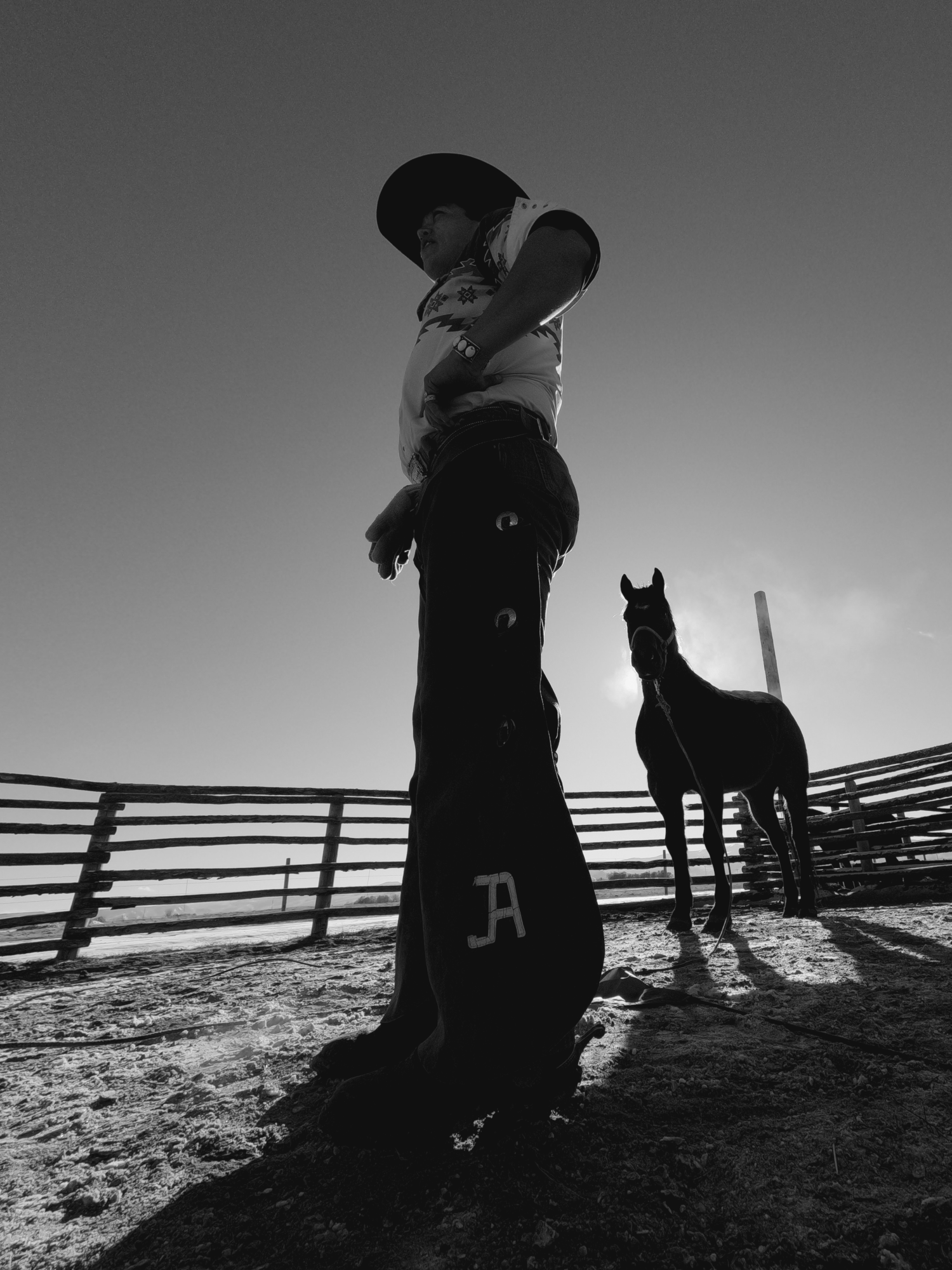 Un cowboy portant un chapeau et des jambières se tient près d'un cheval dans un enclos clôturé sous un ciel clair.