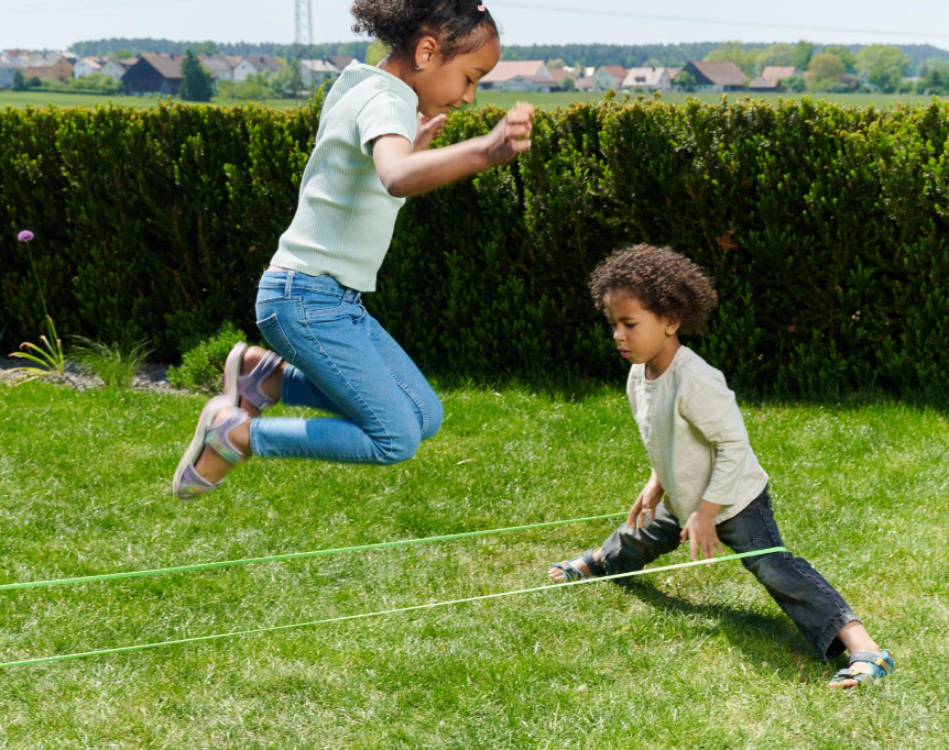 Zwei Kinder spielen Seilspringen im hinteren Garten mit Hecke und Häusern.