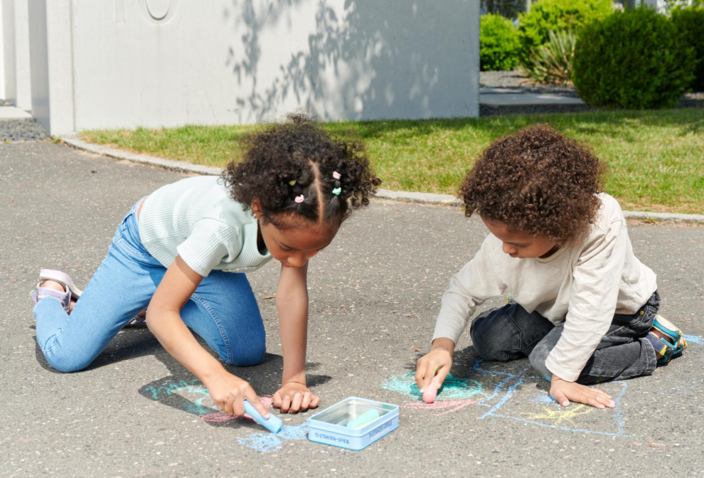 Zwei Kinder malen mit Kreide auf dem Asphalt, umgeben von Grün.