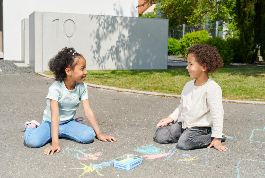 Zwei Kinder malen mit Kreide auf Asphalt, im Hintergrund ein Haus und Bäume.