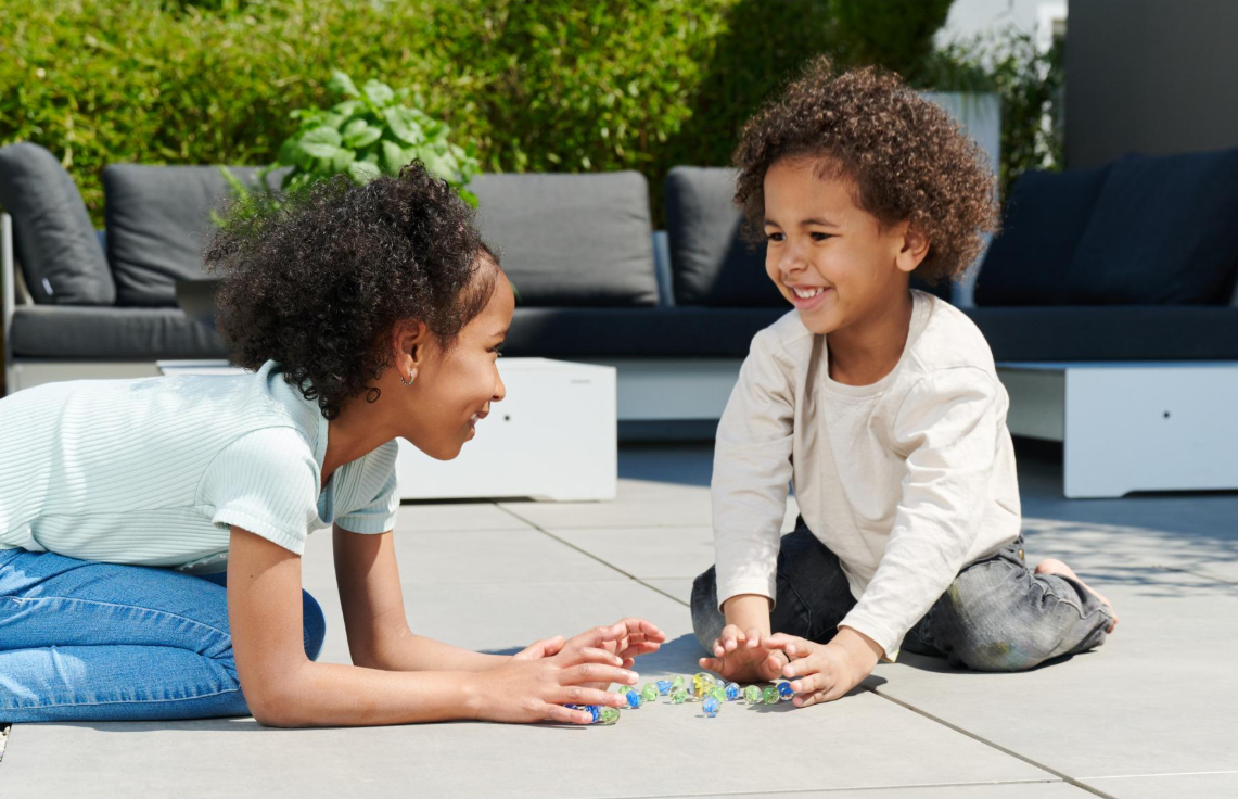 Zwei Kinder spielen draußen mit Murmeln auf einer Terrasse.