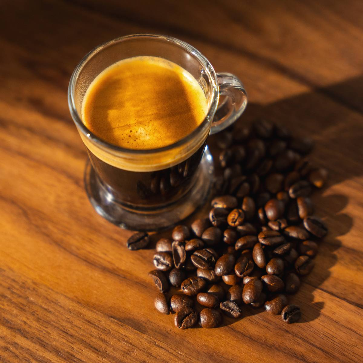 Un vaso de espresso con crema junto a una pila de granos de café tostado sobre madera.