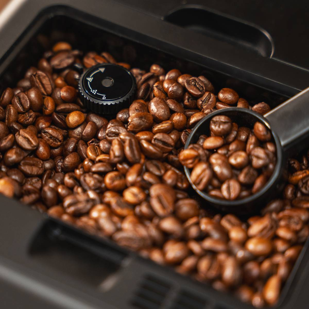 Un recipiente negro lleno de granos de café tostado con una cuchara y un dial de ajuste.