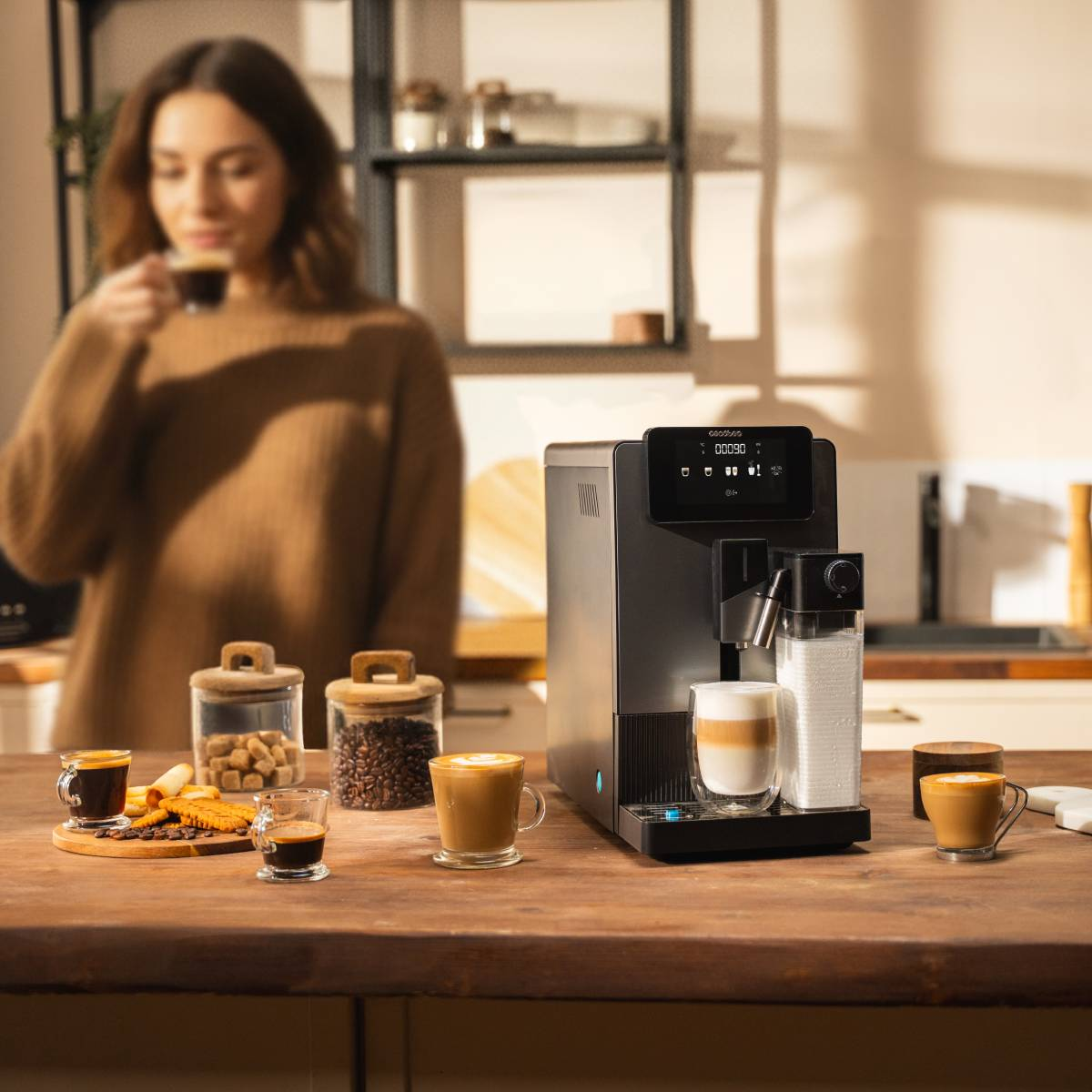 Una mujer bebe café junto a una cafetera con varias tazas y galletas.
