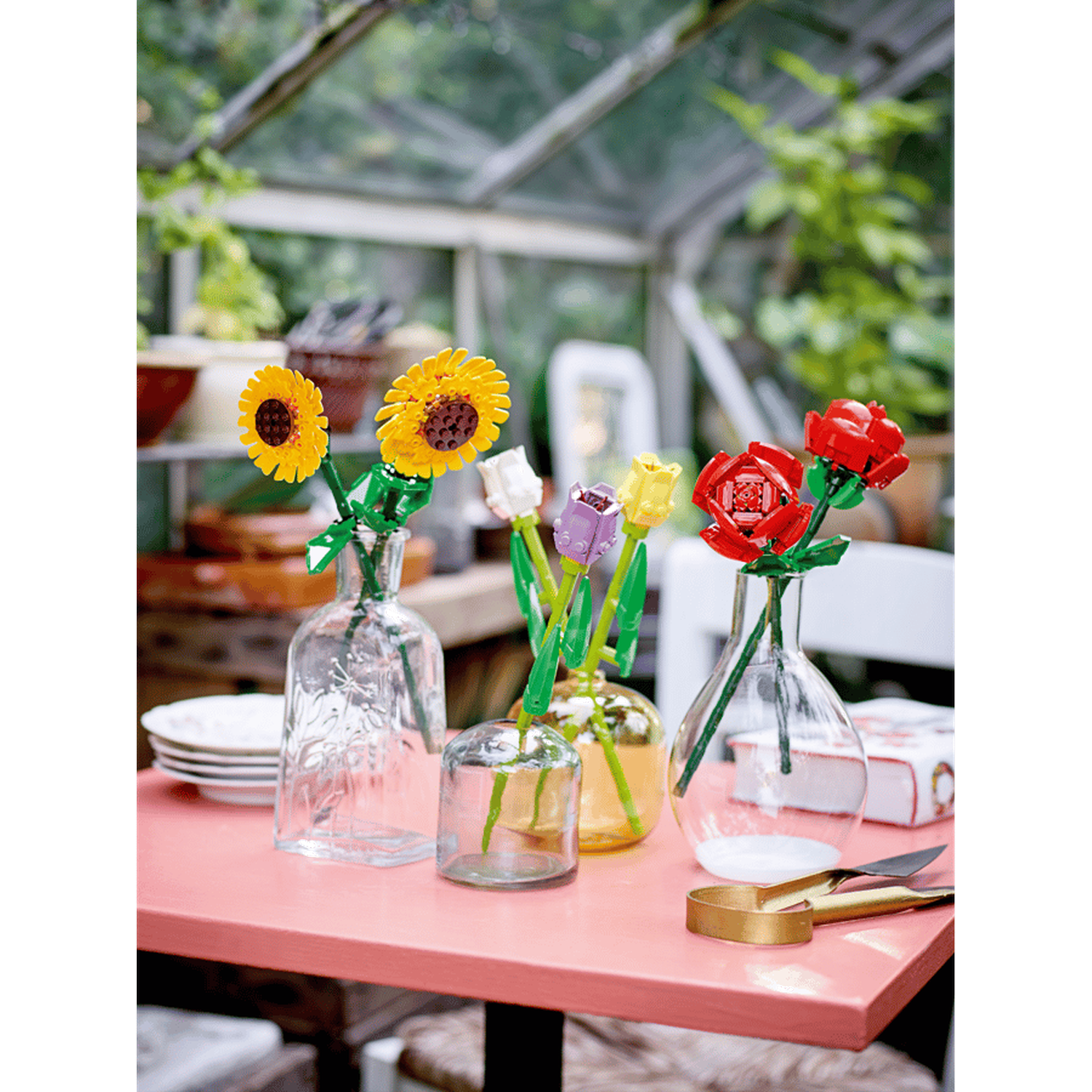 Colorful LEGO flowers in glass vases on a pink table in a greenhouse.