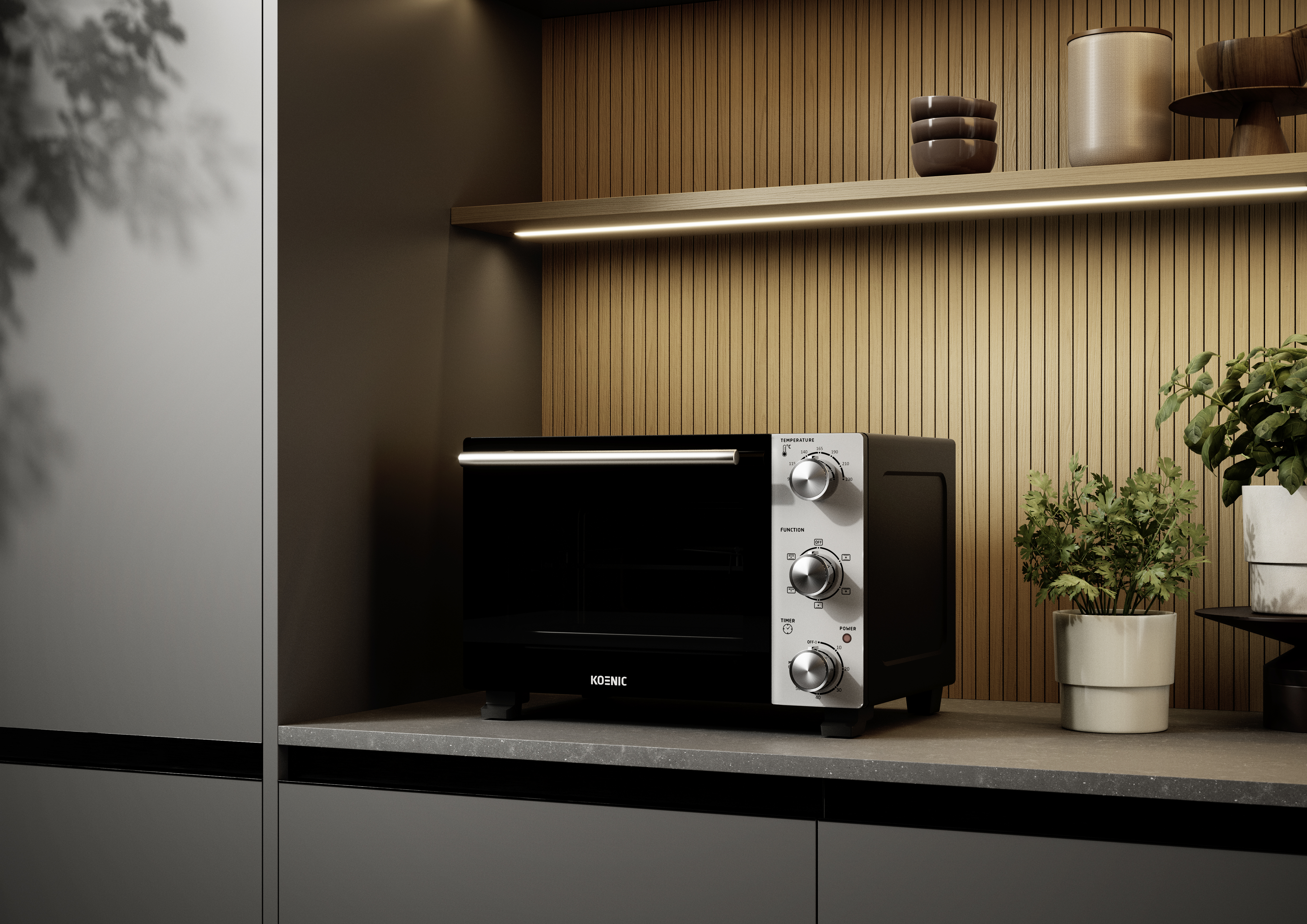 A black KOENIC oven sits on a dark countertop with wood paneling and plants behind it.