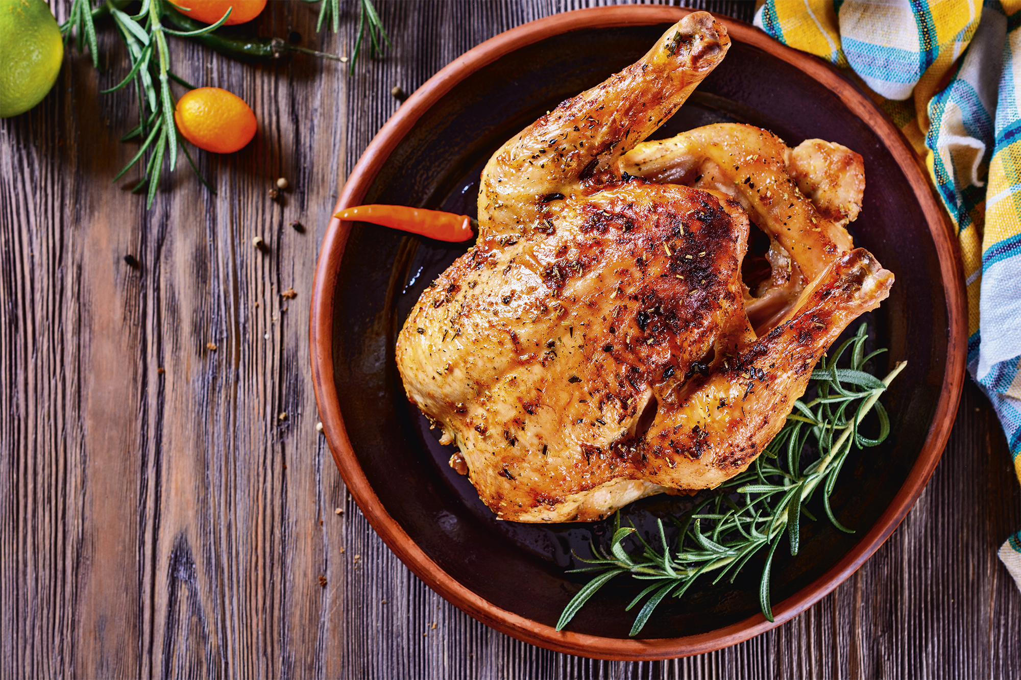 A roasted chicken lies on a brown plate with rosemary and a chili pepper.