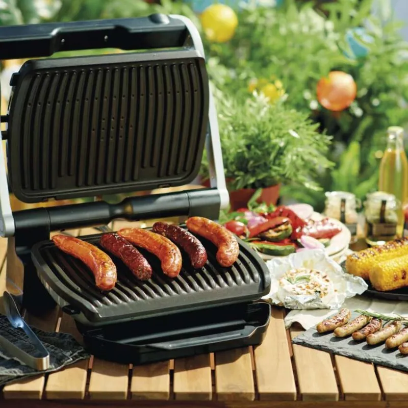 Kolbászok sülnek nyitott grillen, sült zöldségekkel és kukoricával faasztalon.