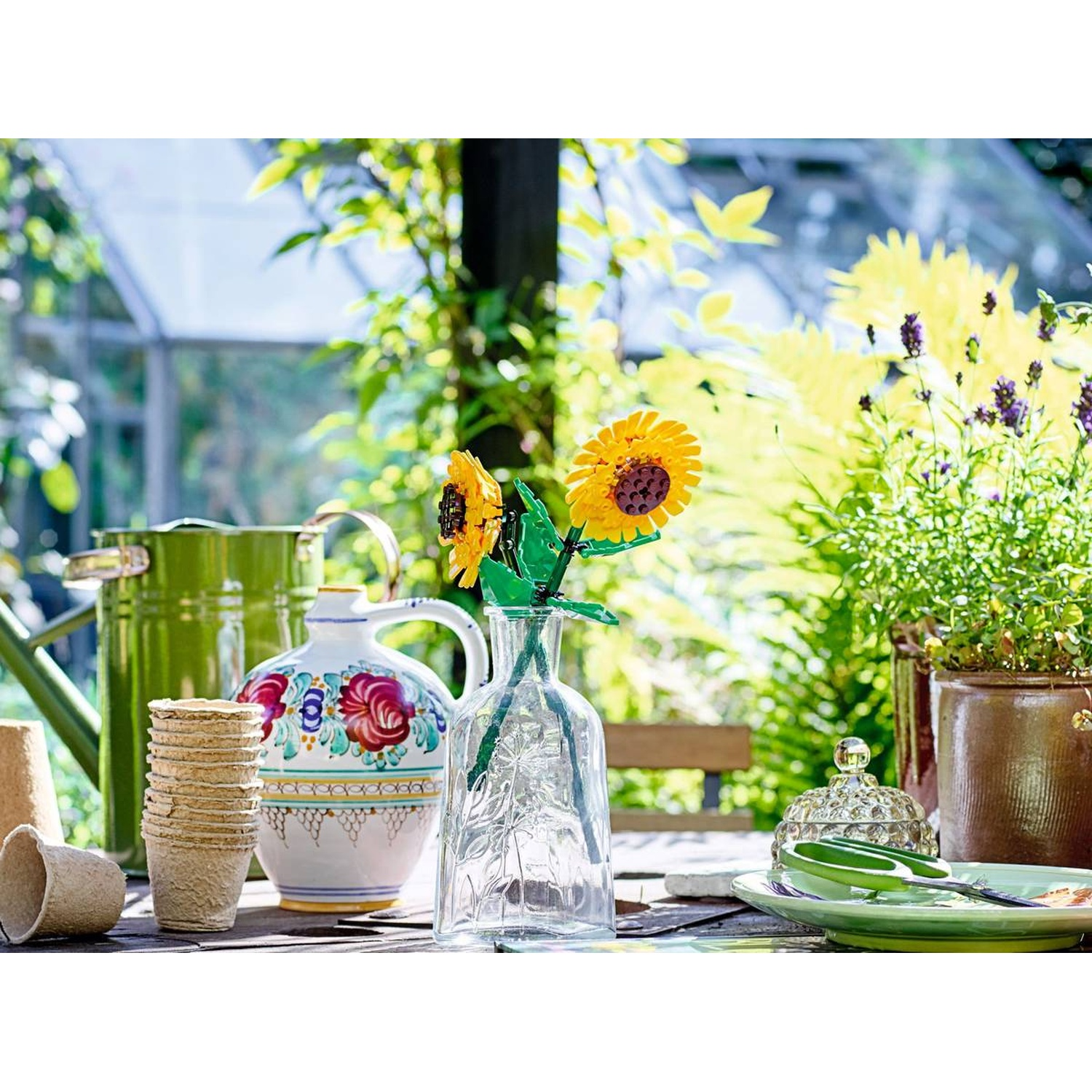 Une table avec des articles de jardin : arrosoir, vase, pots, fleurs, assiette.