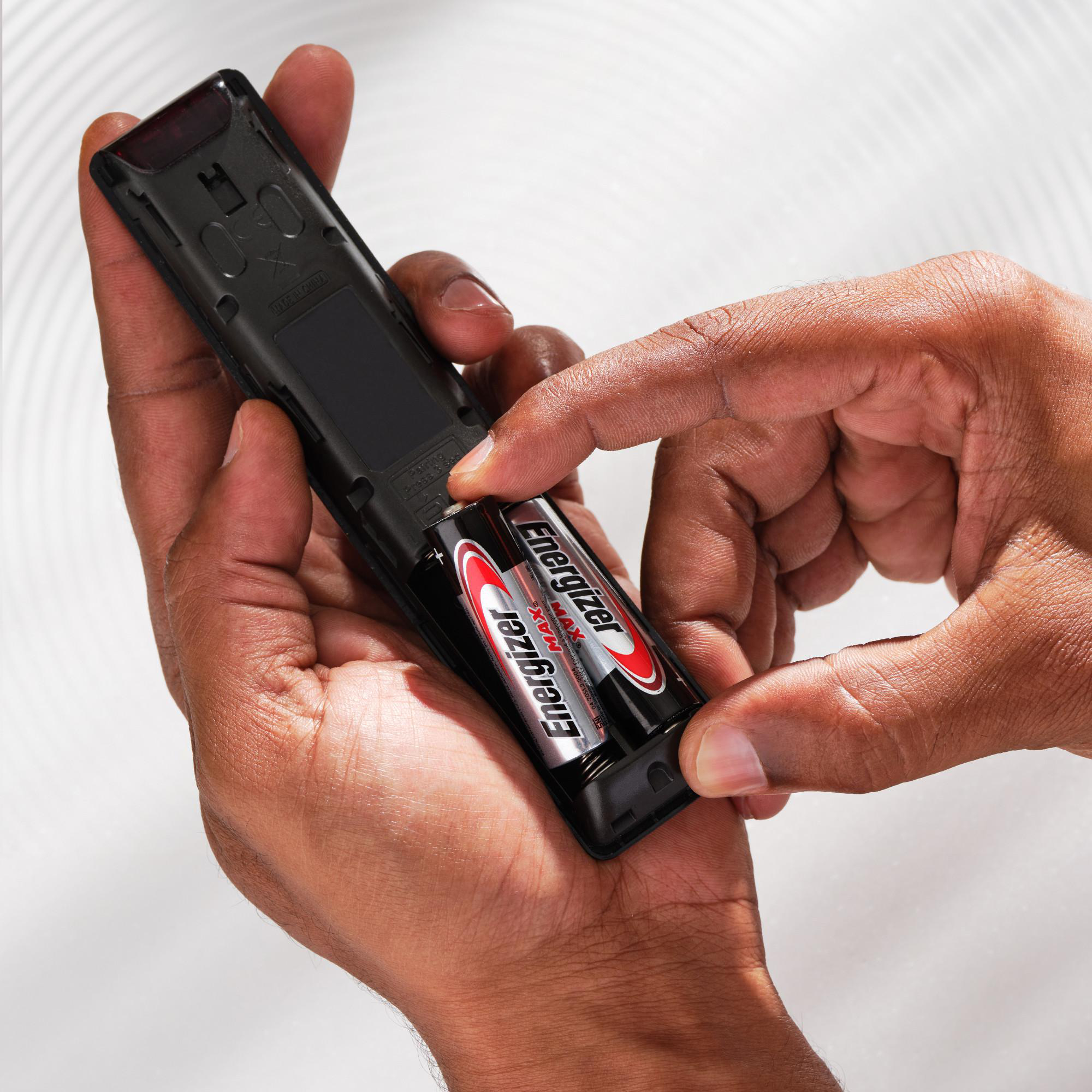 Hände setzen Energizer MAX Batterien in eine schwarze Fernbedienung ein.
