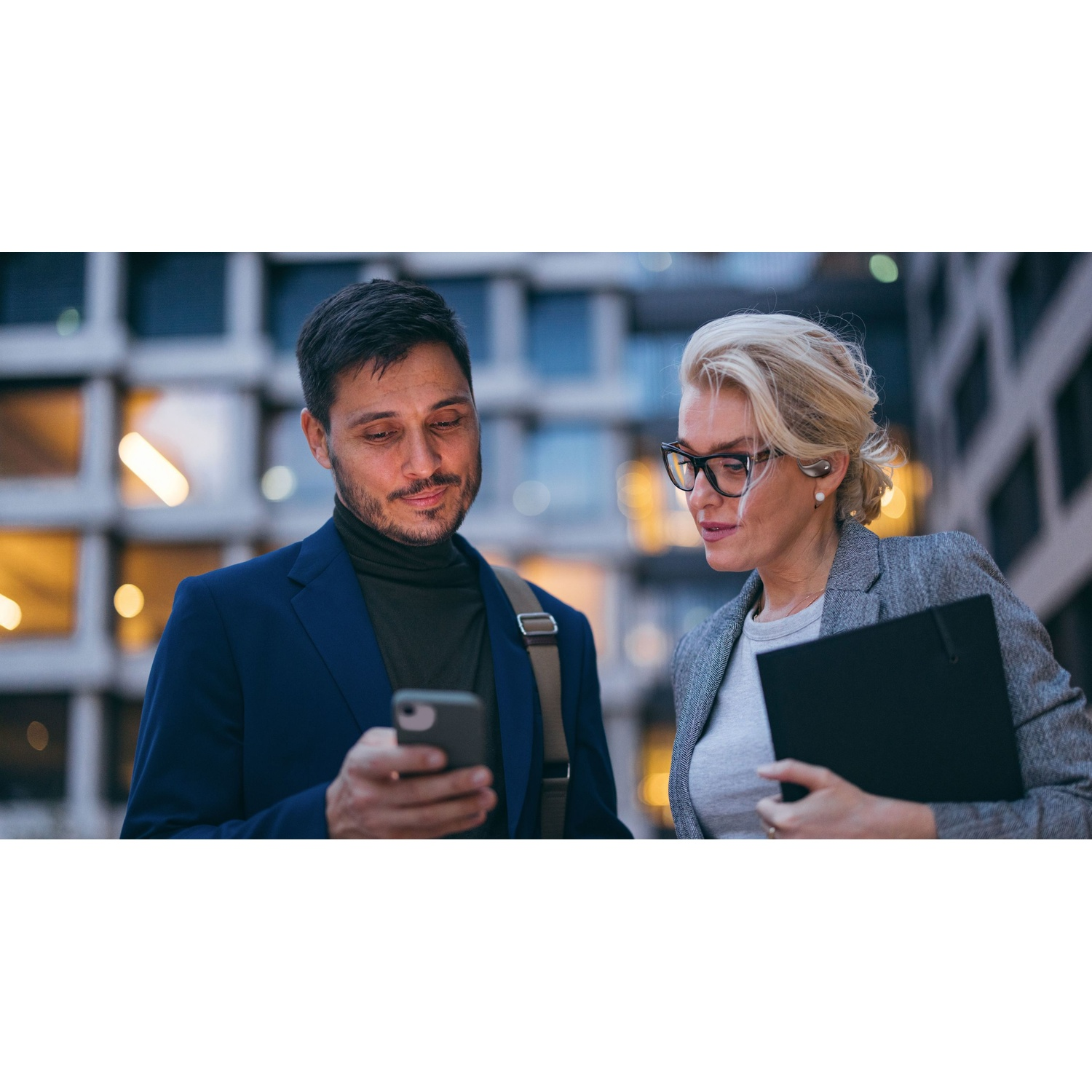 Uomo e donna guardano un telefono. Uomo in abito blu, donna in blazer grigio.