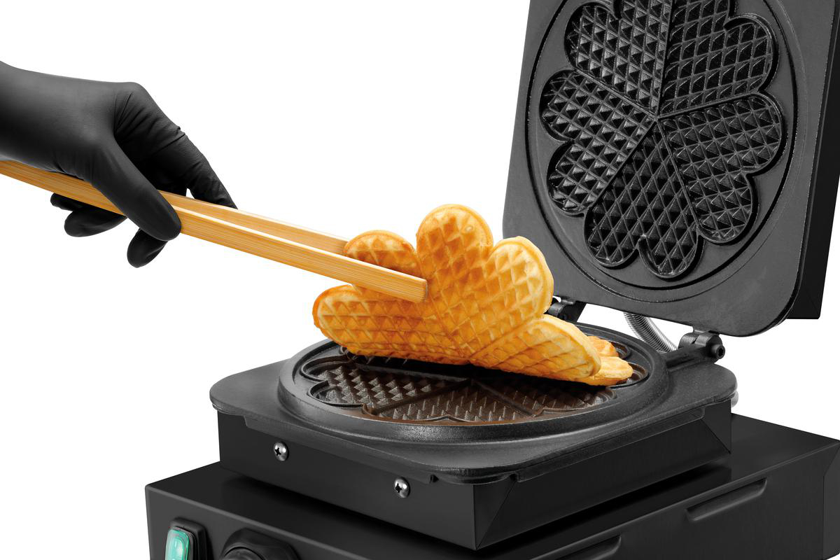 Eine Hand in einem schwarzen Handschuh benutzt Holzgriffe, um herzförmige Waffeln aus einem Waffeleisen zu nehmen.