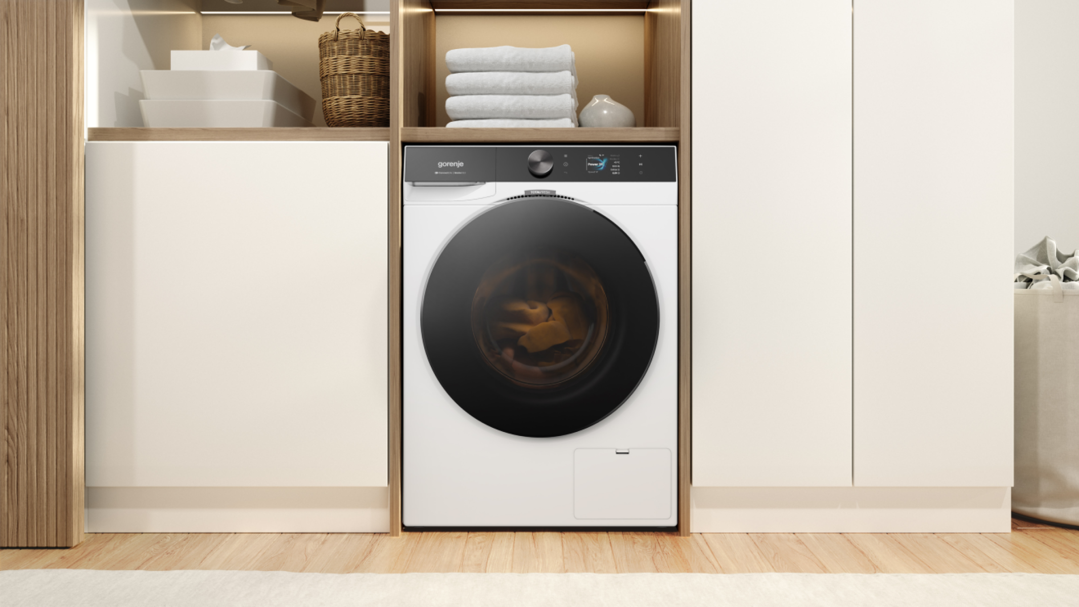 Egy fehér Gorenje mosógép fekete ajtóval egy fa polcon áll. Felül törölközők vannak.