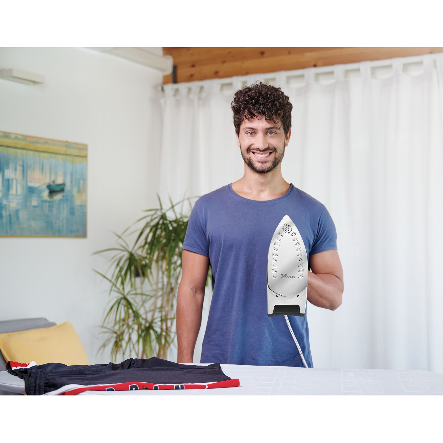 Un uomo con i capelli ricci tiene un ferro da stiro su un asse da stiro con vestiti.