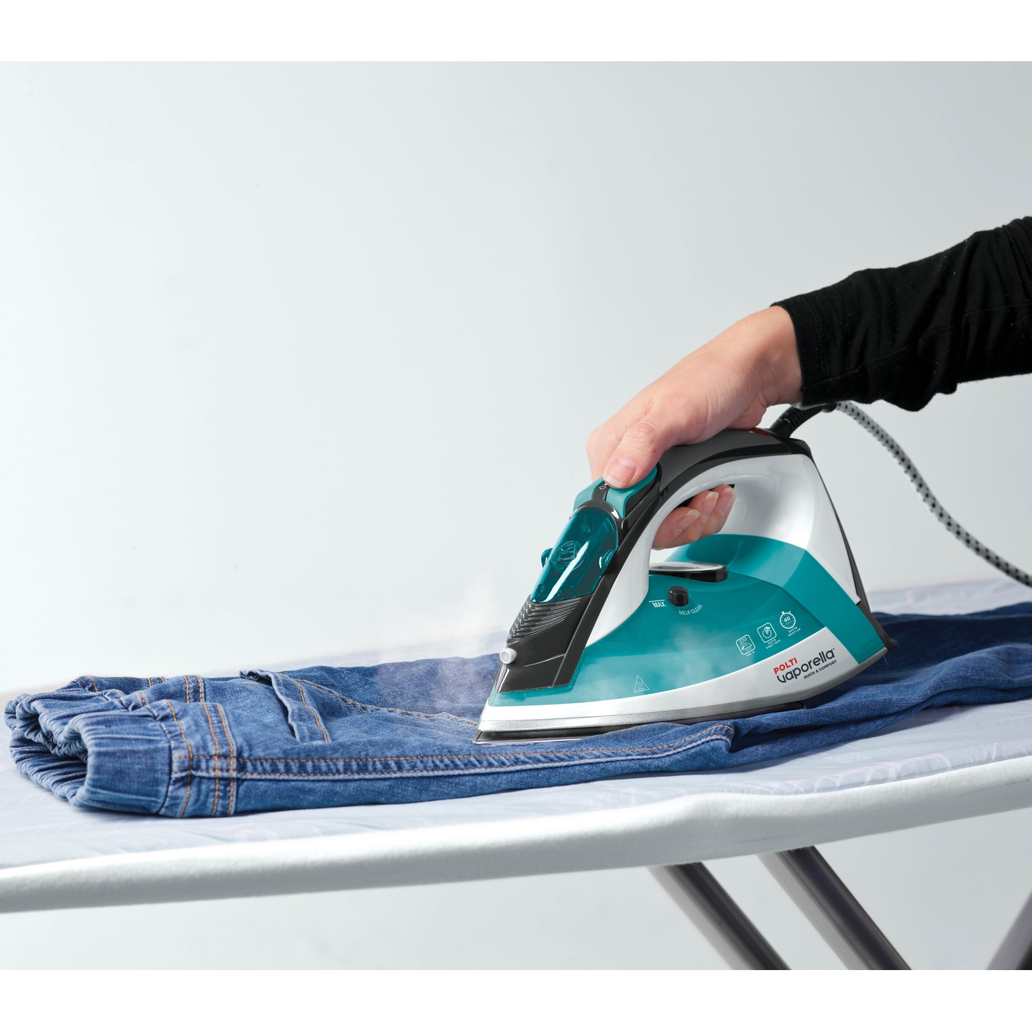 Donna stira jeans blu su asse da stiro bianco, vapore sale dal ferro.