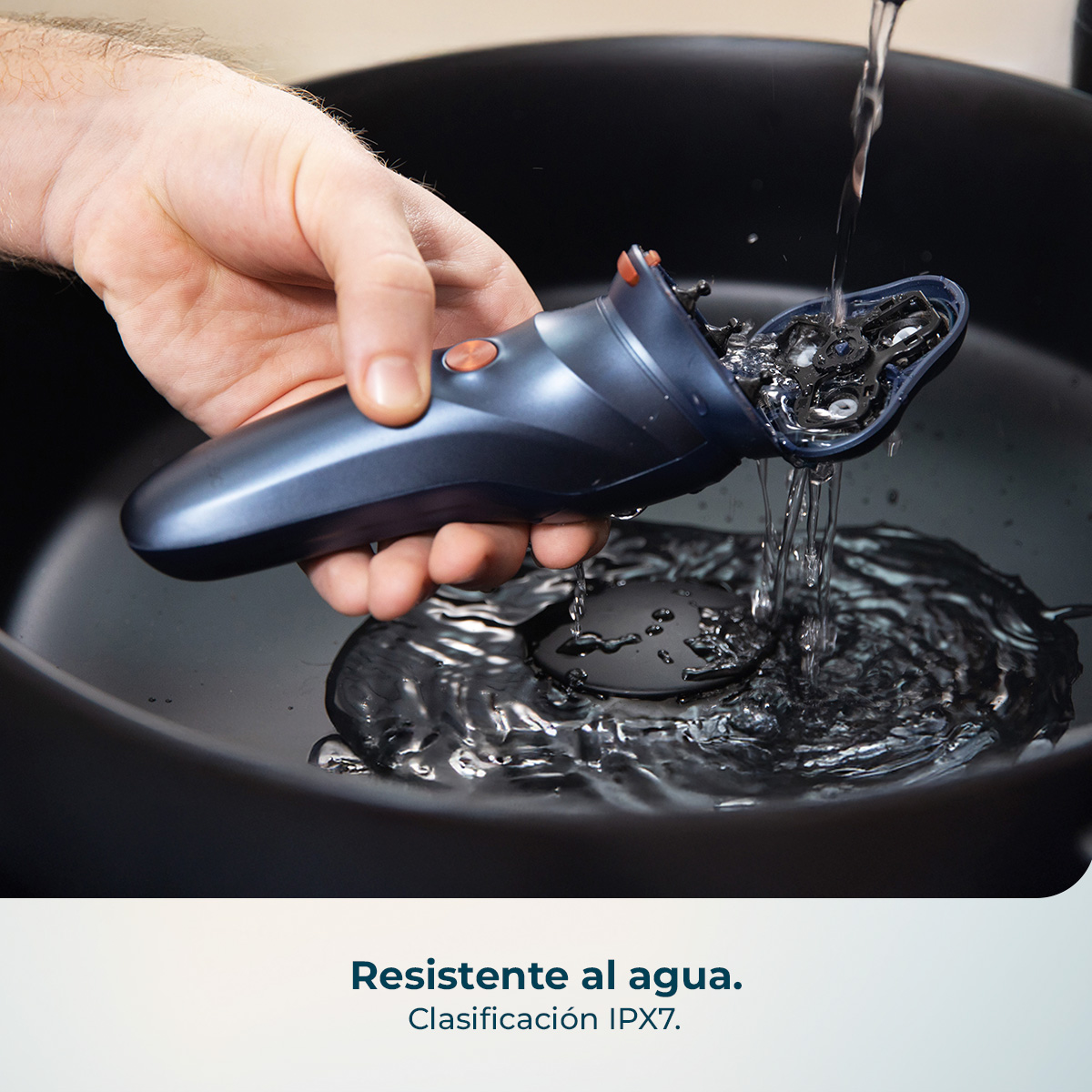 Una mano sostiene una afeitadora eléctrica oscura bajo agua corriente en un cuenco negro.