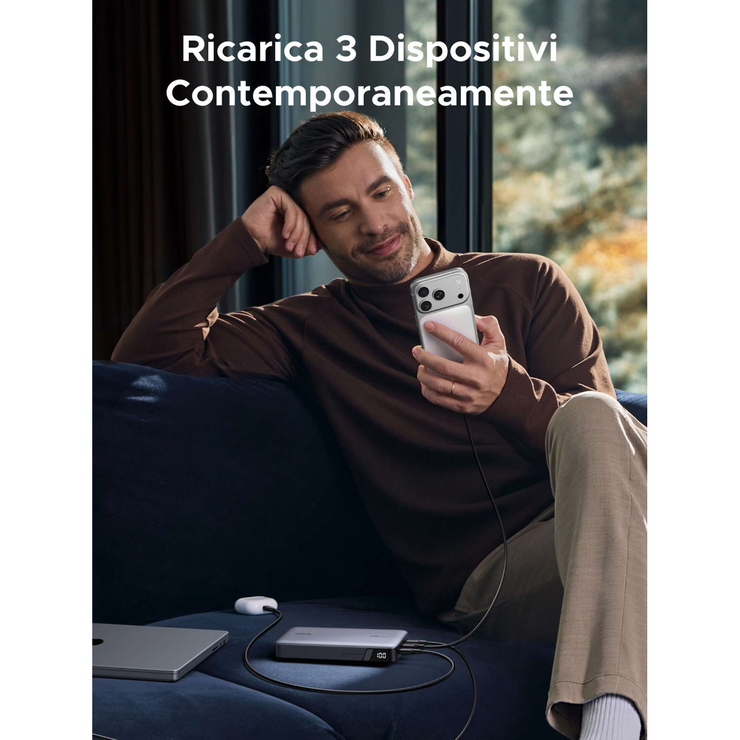 Uomo su divano con telefono, laptop, power bank e auricolari, carica dispositivi.