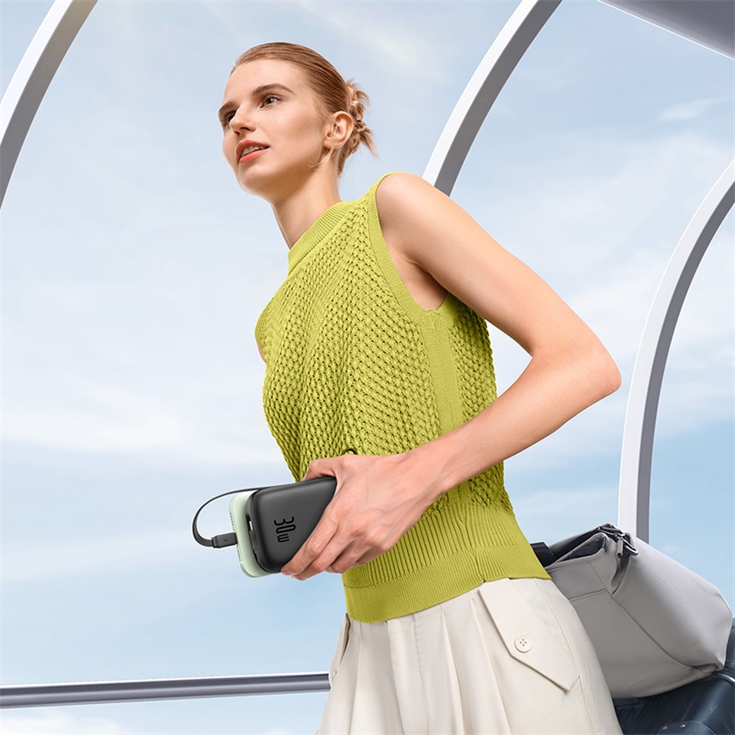 Mujer con top verde lima sostiene power bank negra y dispositivo verde claro. Bolso gris a la derecha.