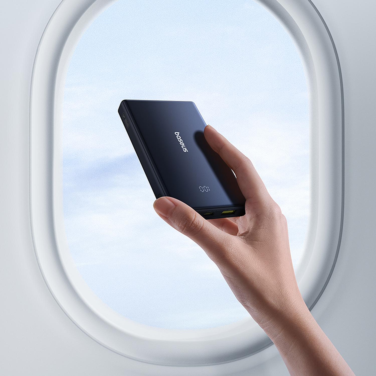 Una mano sostiene un cargador Baseus negro frente a la ventana de un avión.