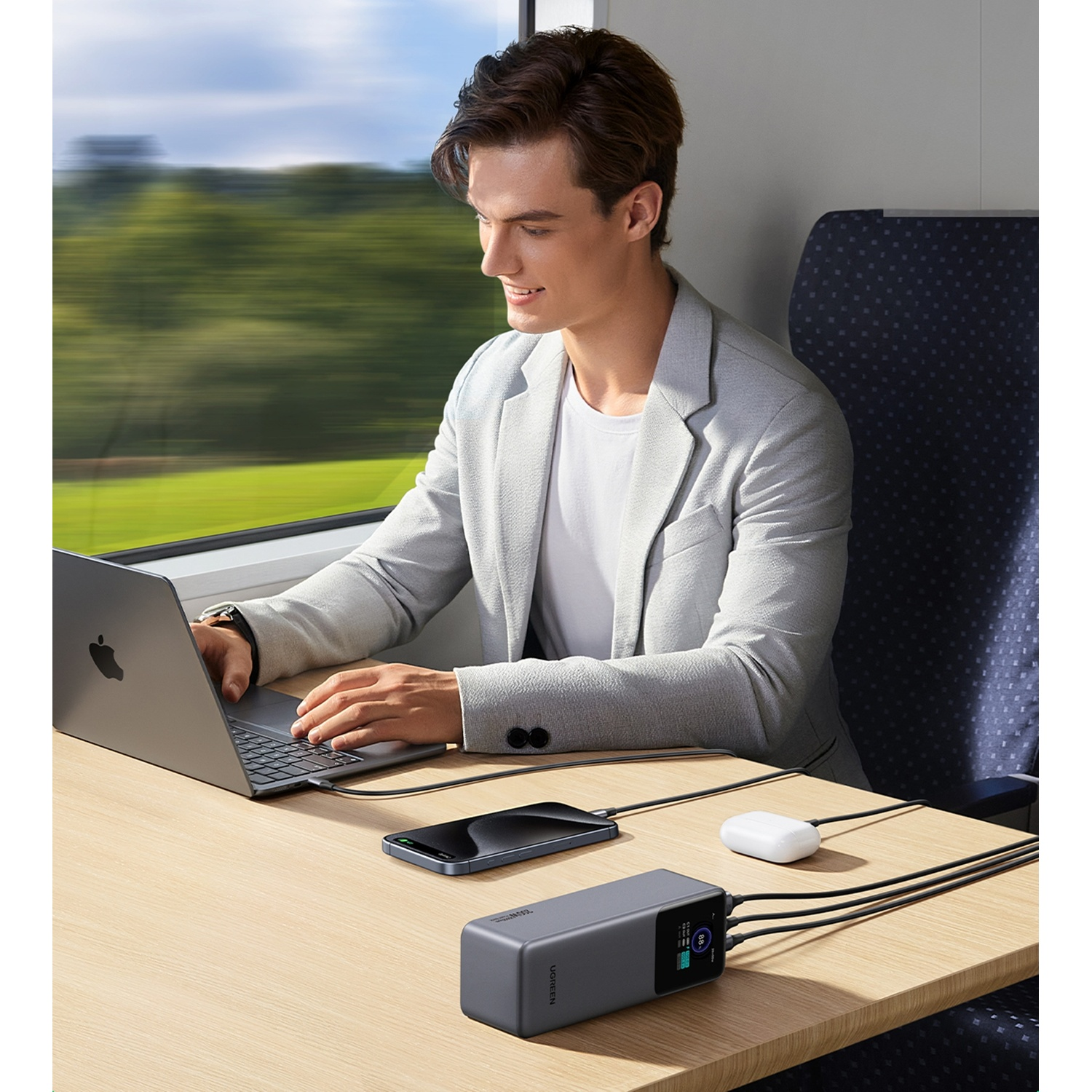 Uomo su un treno lavora al laptop, con telefono, power bank e AirPods nelle vicinanze.