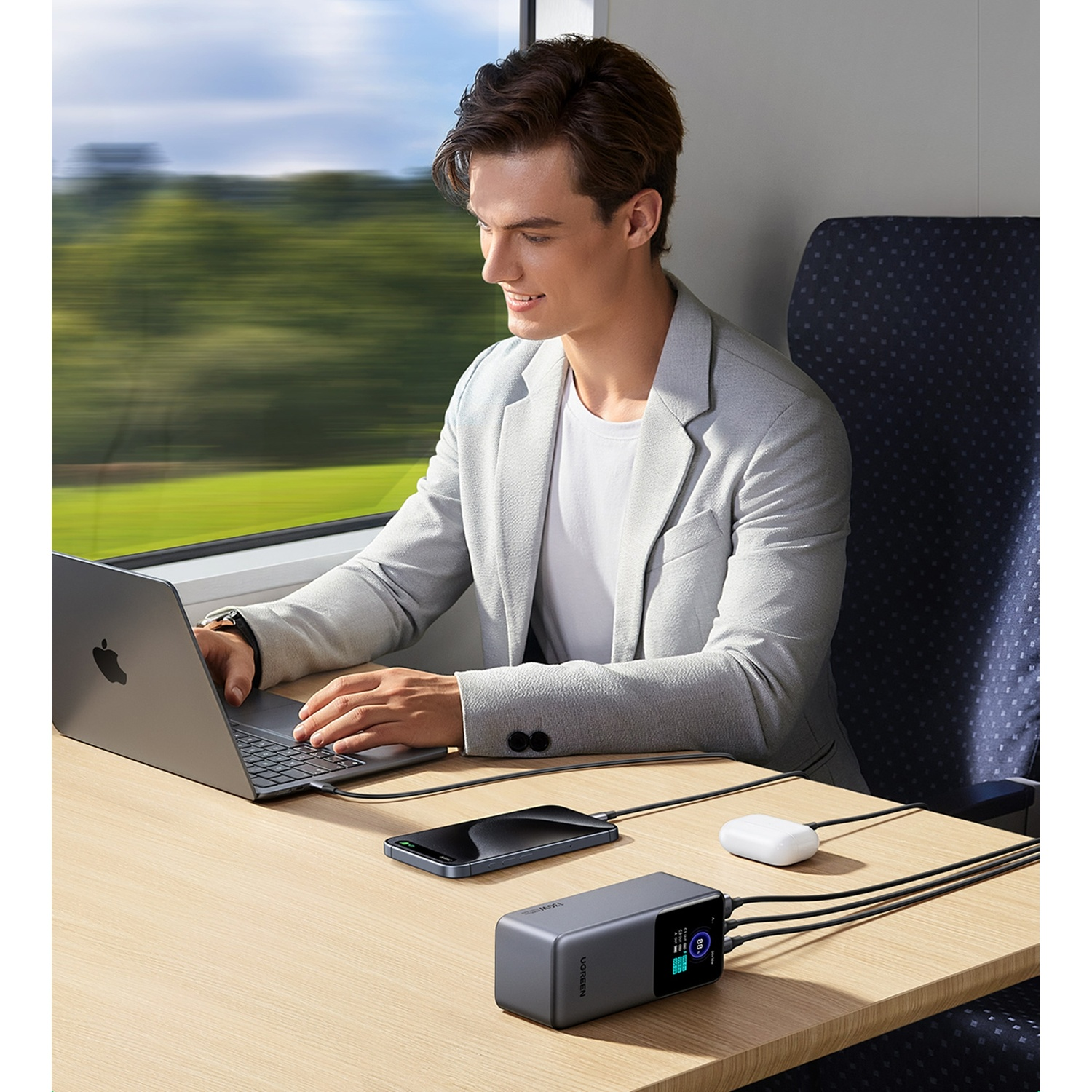 Uomo in treno usa laptop, telefono e caricabatterie. Paesaggio verde fuori.