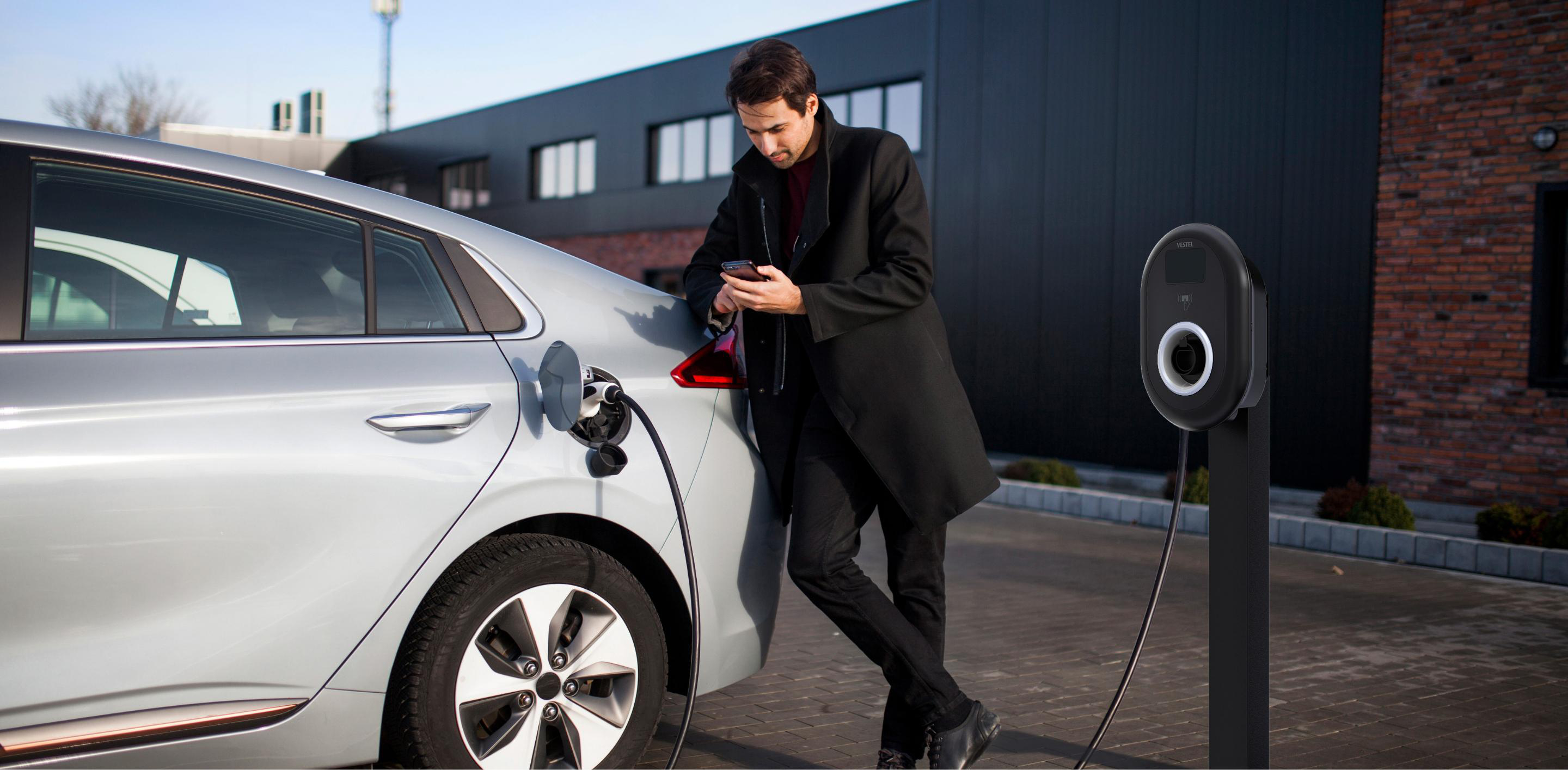 Mann mit Handy lehnt an einem silbernen Elektroauto, das an Ladestation lädt.
