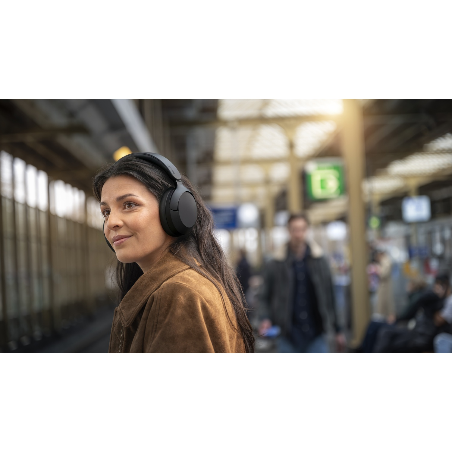 Donna con cuffie alla stazione, giacca marrone, sfondo sfocato con persone.