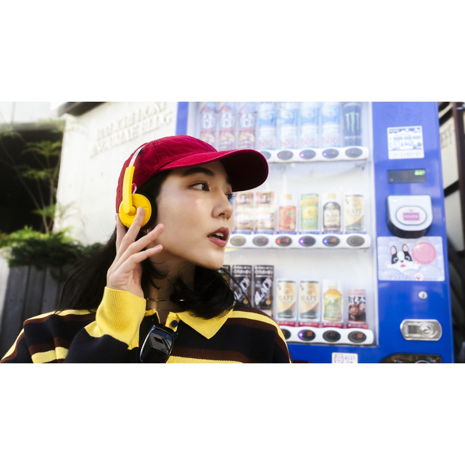 Una donna con cappellino rosso e cuffie gialle davanti a un distributore automatico.