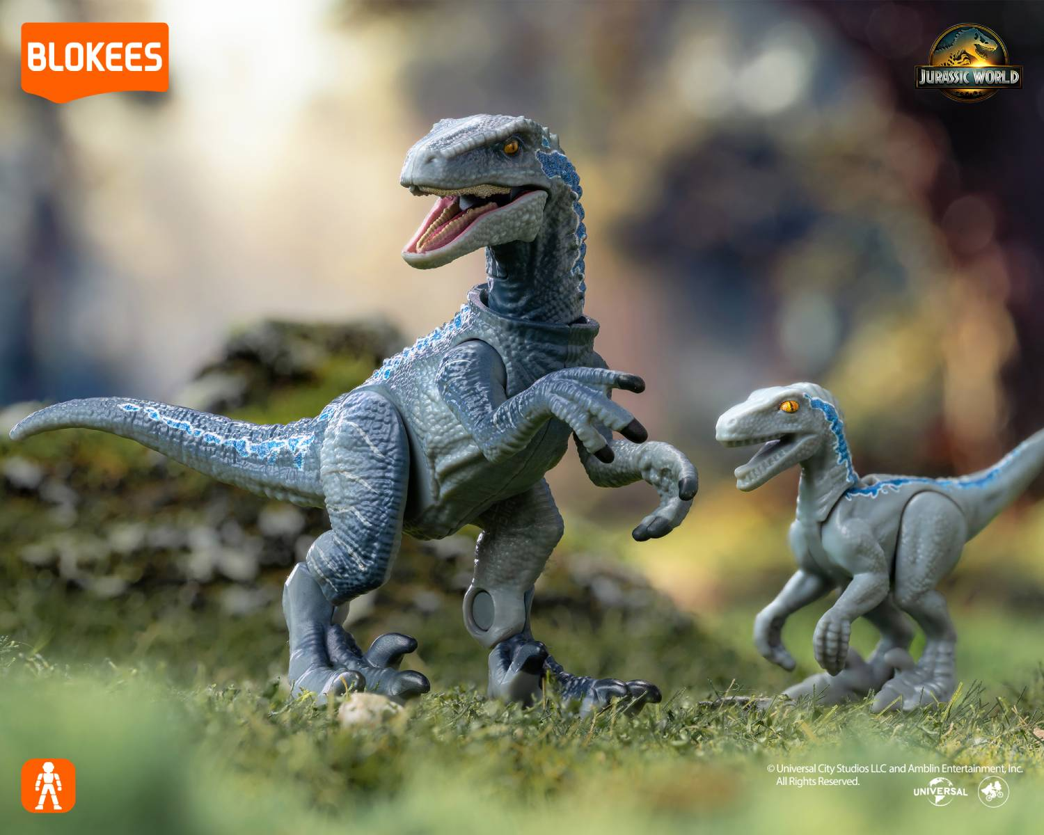Dos juguetes de velociraptor azules en hierba verde, uno grande, uno pequeño. Logo Jurassic World.