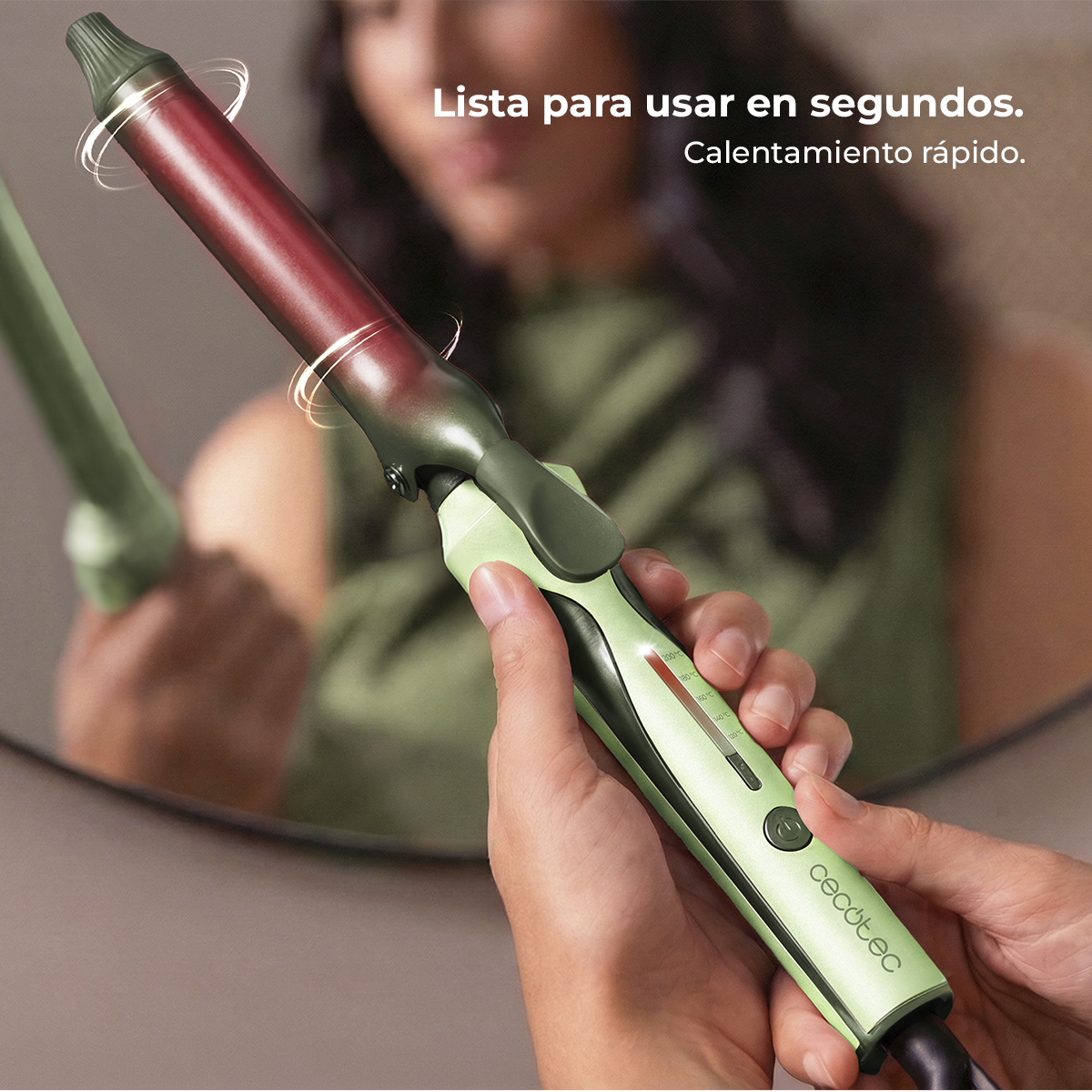 Una persona sostiene un rizador verde con barril rojo, reflejado en un espejo.