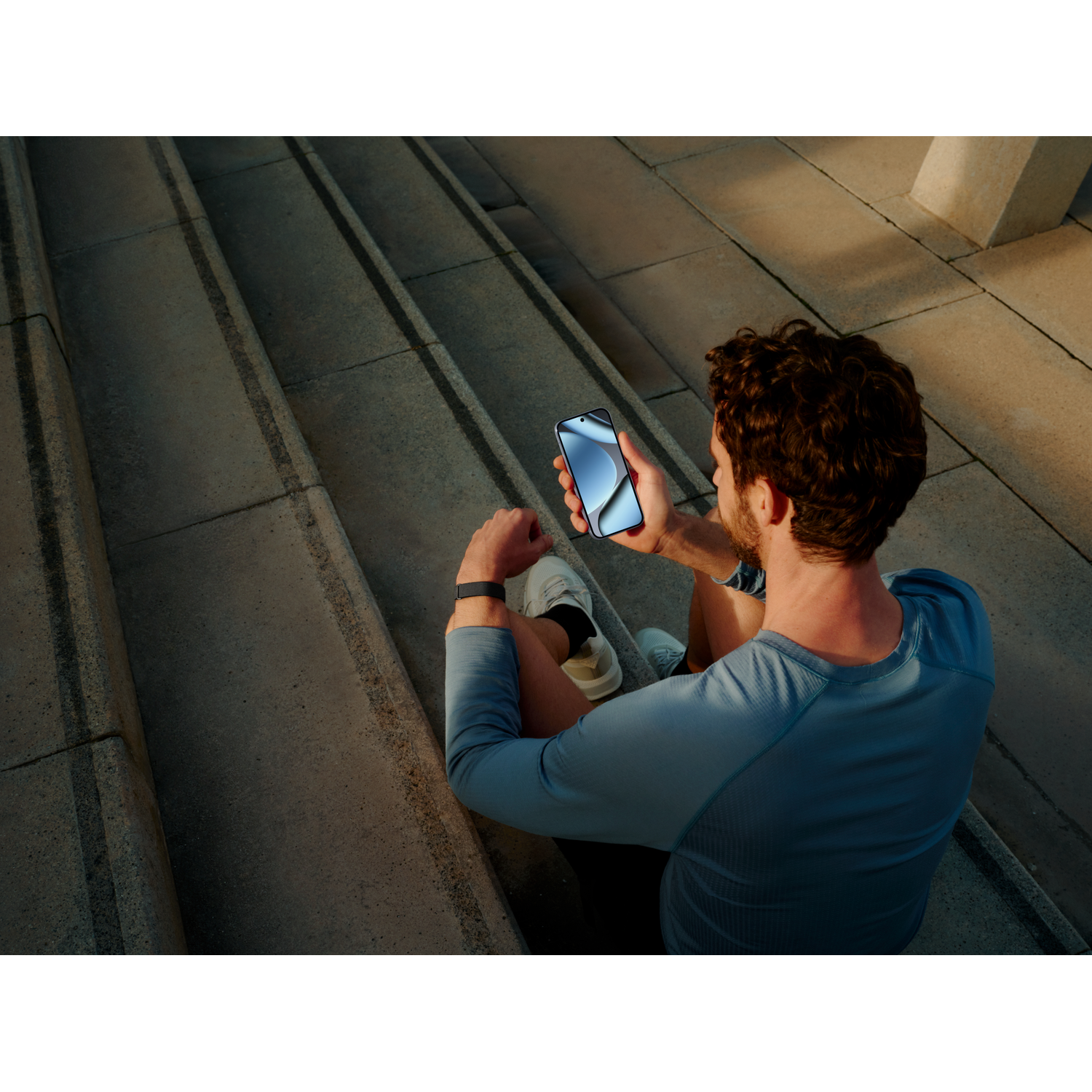Man in blue shirt sits on concrete steps holding a smartphone.