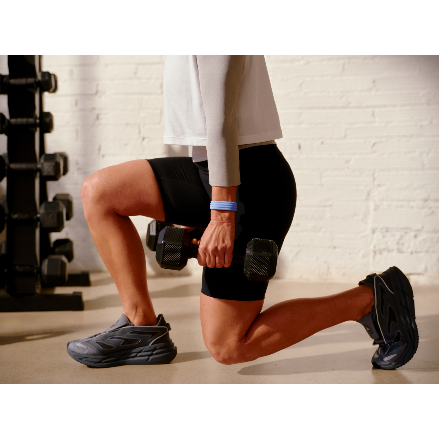 Persoon doet lunge met dumbbell, gewichten op achtergrond. Korte donkere broek, grijs shirt.