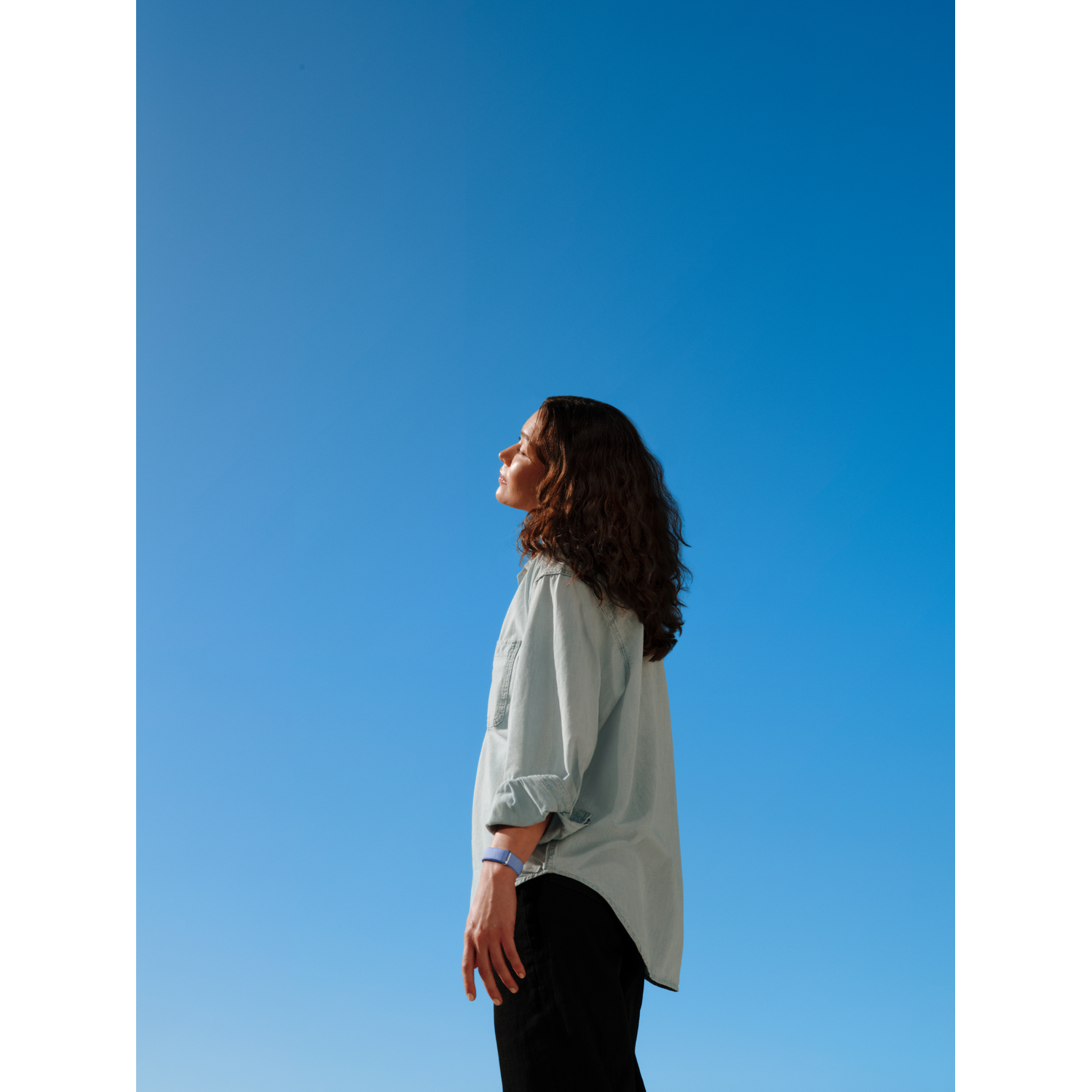 Vrouw kijkt omhoog naar helderblauwe lucht, draagt lichtblauw shirt en zwarte broek.