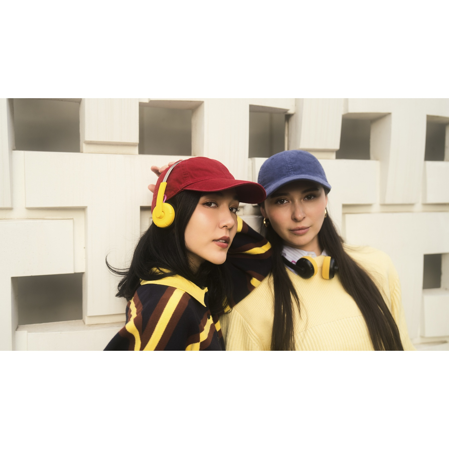 Due donne indossano cuffie e cappellini, posando davanti a un muro bianco.