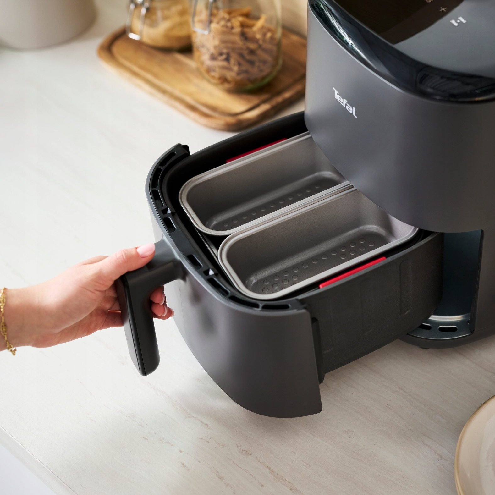 Ręka wyciąga szufladę czarnej frytkownicy beztłuszczowej Tefal z dwoma foremkami do pieczenia.