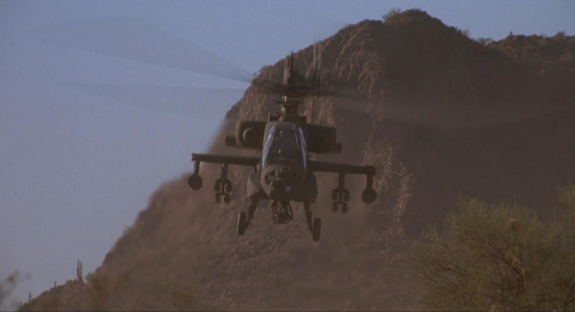 Ein dunkler Kampfhubschrauber fliegt vor einer staubigen Bergkulisse.