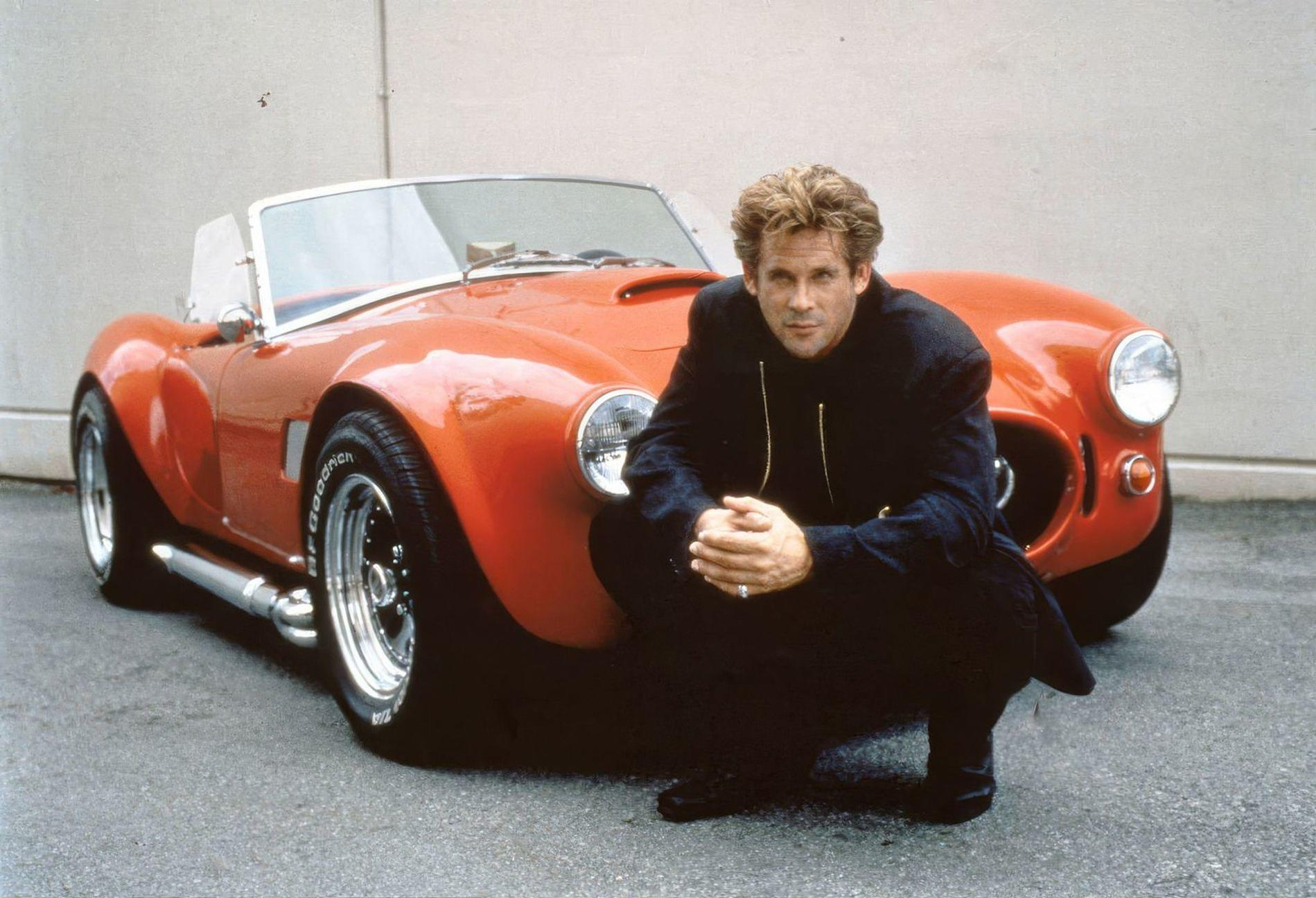 Ein Mann in Schwarz kniet neben einem roten Oldtimer-Sportwagen.
