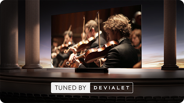 Orkiestra gra na skrzypcach przed kolumnami na scenie z napisem "TUNED BY DEVIALET".