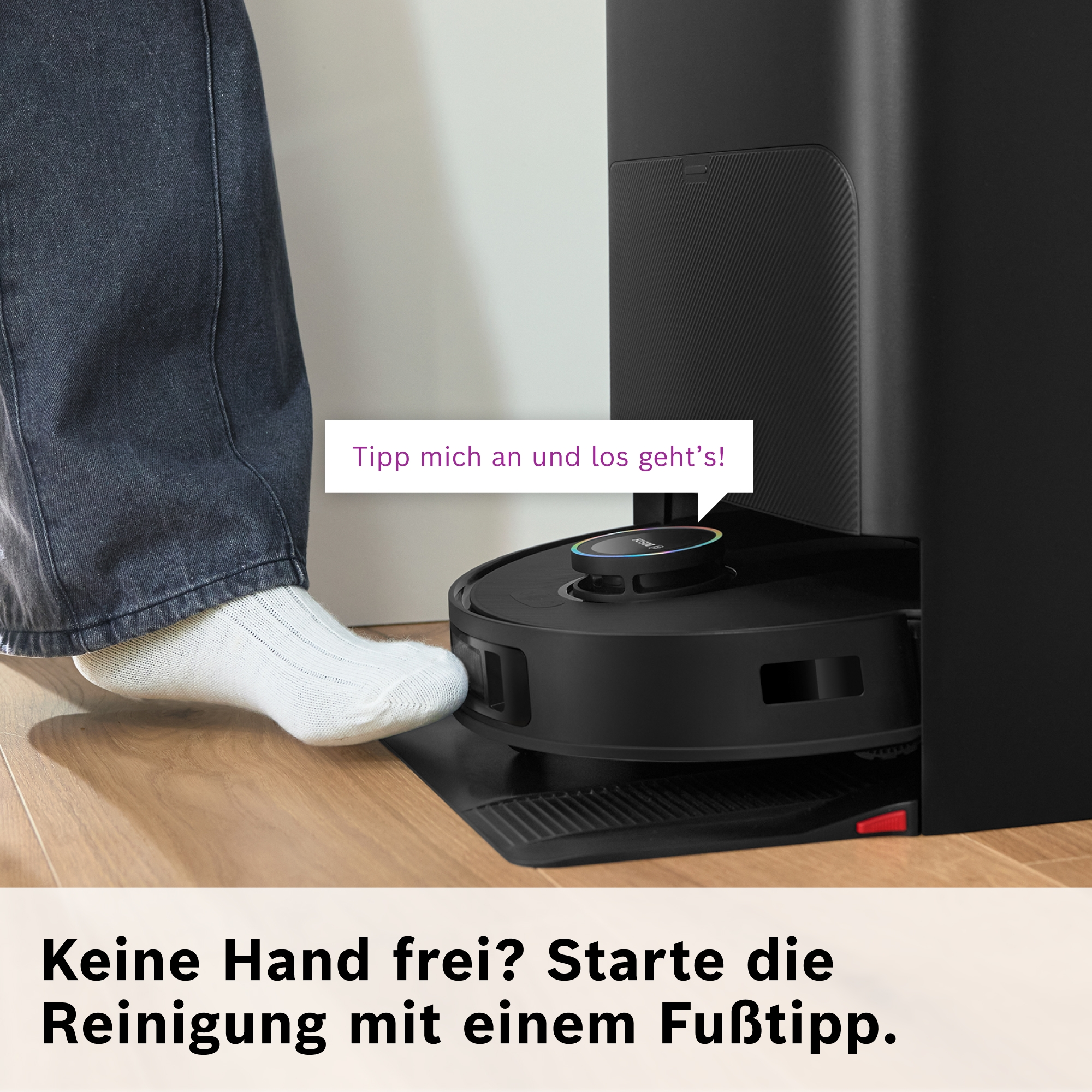 Fuß startet Saugroboter. Schwarzer Roboter mit Lichtring, Person in Jeans und Socken.
