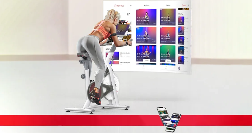 Kobieta na rowerze stacjonarnym, ekran pokazuje filmy treningowe, telefony poniżej.